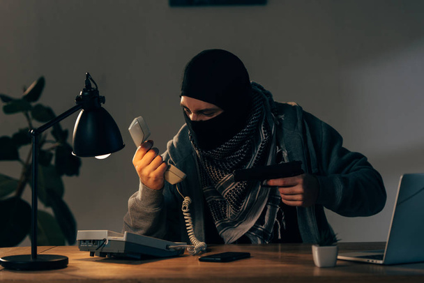 Verbrecher mit schwarzer Maske hält Pistole in der Hand und schaut auf Handy - Foto, Bild