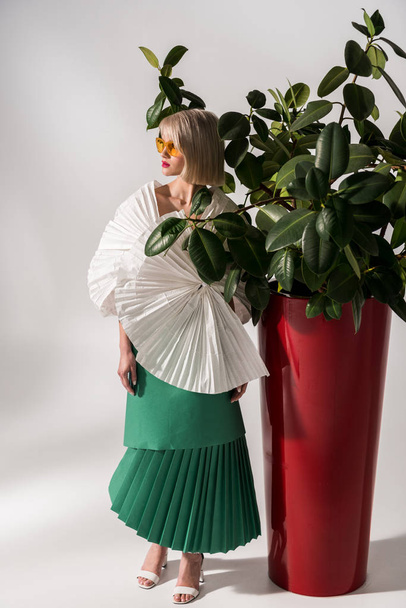 красива стильна дівчина в сонцезахисних окулярах і паперовому одязі позує біля рослини на сірому
 - Фото, зображення