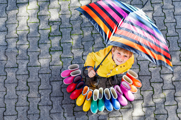 Niño pequeño y grupo de botas de lluvia de colores. Niño rubio de pie bajo el paraguas
. - Foto, Imagen