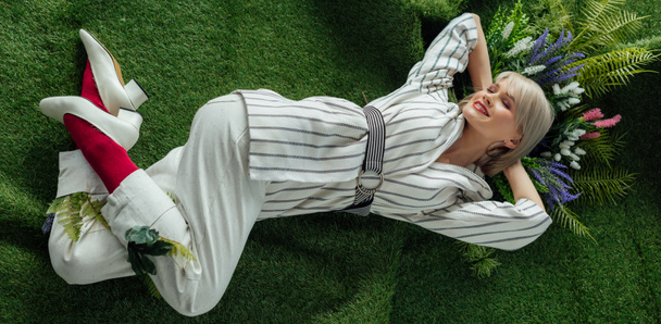 panoramic shot of beautiful stylish girl smiling while lying on artificial grass with flowers  - Photo, Image