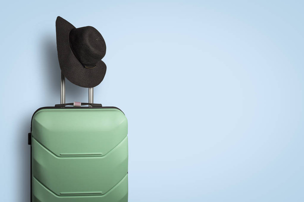 Plastic suitcase on wheels and a hat with a wide brim on the handle on a blue background. Travel concept, vacation trip, visit to relatives - Foto, Imagem