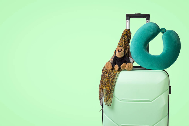 Plastic suitcase with wheels, soft toy, travel pillow, neck scarf, green background. Concept of travel on a summer vacation, a visit to relatives, a trip to the camp - Photo, Image
