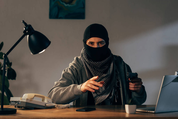 Terrorist in mask sitting at table with lamp and holding gun - Photo, Image
