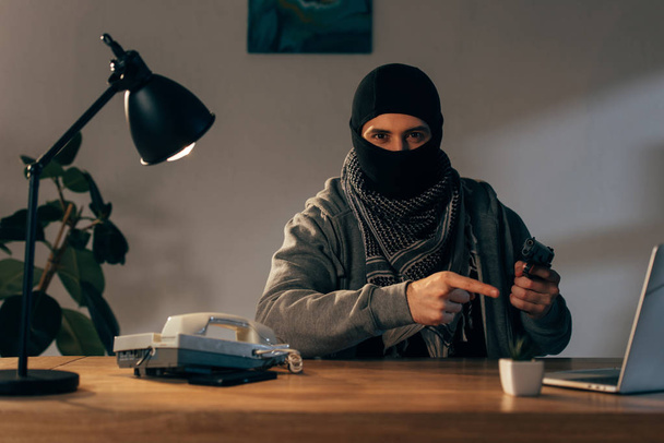 Terroristi mustassa naamiossa istuu pöydässä ja osoittaa aseella sormella
 - Valokuva, kuva