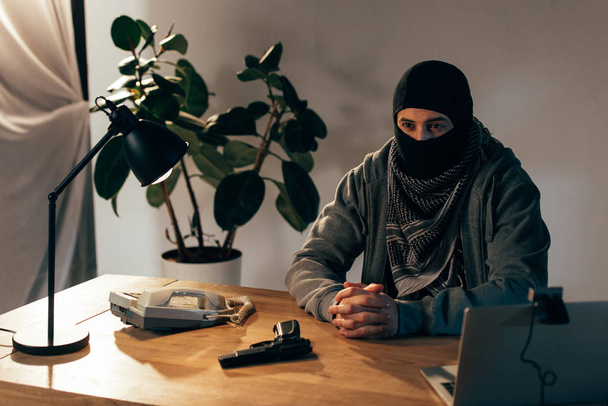 Nachdenklicher Verbrecher in schwarzer Maske sitzt mit verschränkten Fingern am Tisch - Foto, Bild