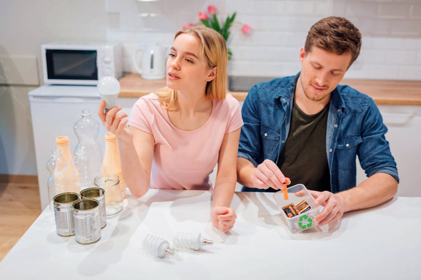 Recycling, reuse. Young couple sorting batteries, light bulbs into small containers with recycling symbol while sitting at kitchen - Foto, Imagem
