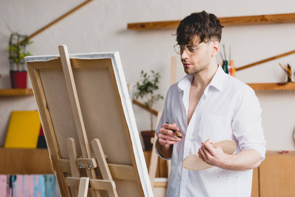 handsome artist in glasses standing at easel with canvas in painting studio - Foto, imagen