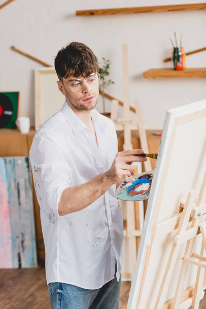 handsome artist in white shirt and glasses painting on canvas in gallery - Photo, Image