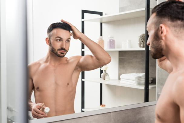 hombre guapo sin camisa tocando el cabello y sosteniendo el cepillo de dientes con pasta de dientes mientras mira el espejo en el baño
  - Foto, Imagen