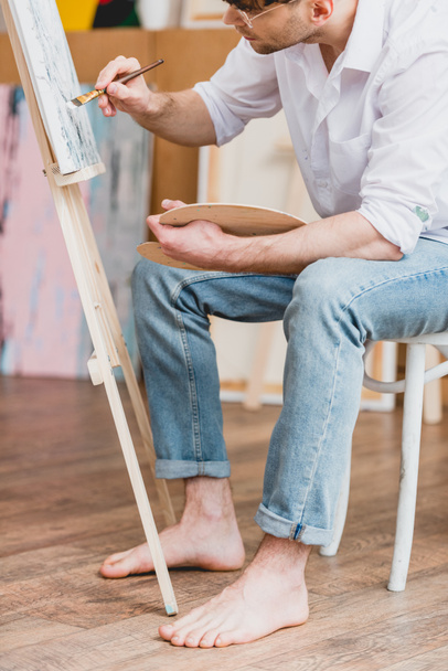 partial view of barefoot artist in white shirt and blue jeans painting on canvas - Photo, Image