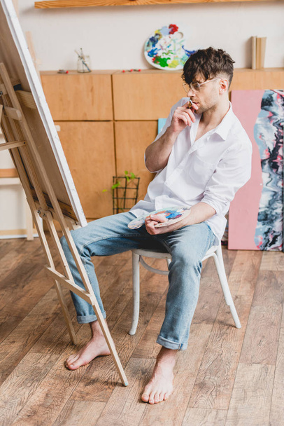 красивый художник в белой рубашке и синих джинсах, смотрящий на холст на мольберте
 - Фото, изображение