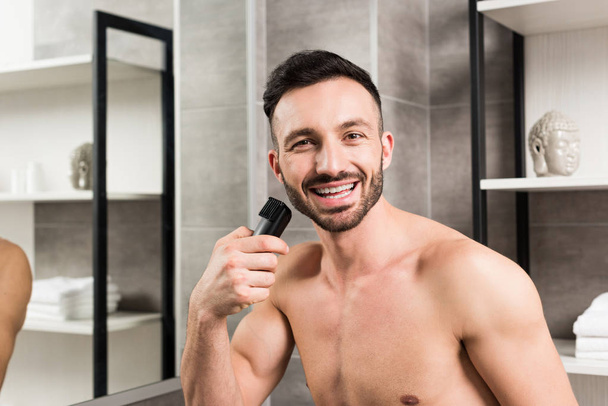 fröhlicher, hemdsloser Mann hält Trimmer im Badezimmer in der Nähe des Gesichts  - Foto, Bild
