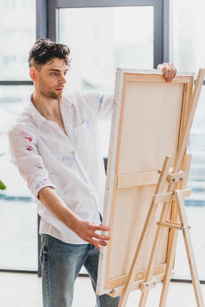 handsome artist in white shirt fixing canvas on easel in painting studio - Foto, afbeelding