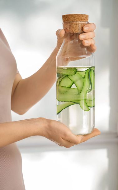 Mujer sosteniendo botella de agua de pepino fresco sobre fondo claro
 - Foto, Imagen