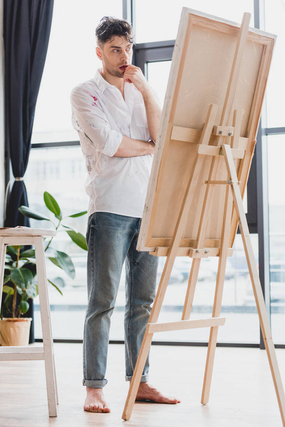 Nachdenklicher Künstler in weißem Hemd und blauer Jeans steht an Staffelei in Galerie - Foto, Bild