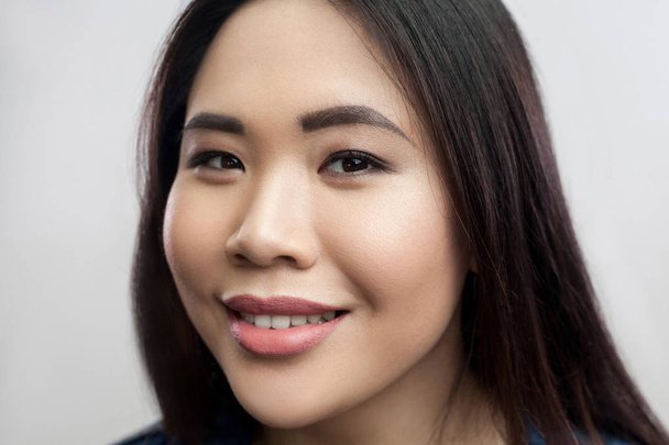 muotokuva iloinen kaunis brunette aasialainen nainen meikki ja suorat tummat hiukset katselee kameraa hammas hymy vaaleanharmaa tausta
  - Valokuva, kuva
