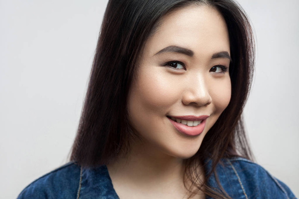 portrait of glad beautiful brunette asian woman with makeup and straight dark hair looking at camera with toothy smile on light grey background   - Φωτογραφία, εικόνα