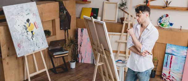 πανοραμικό στιγμιότυπο όμορφος καλλιτέχνη που στέκεται στο καβαλέτο στο εργαστήρι ζωγραφικής - Φωτογραφία, εικόνα