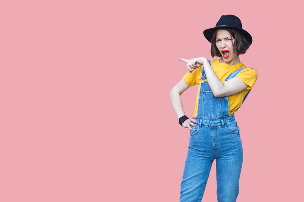 Portrait of amazed beautiful woman in yellow t-shirt and blue denim overalls with makeup and black hat standing with opened mouth and pointing aside on pink background   - Fotografie, Obrázek