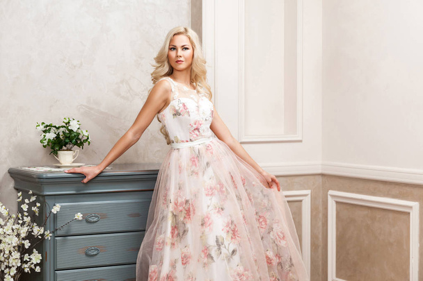 beautiful blonde woman in pastel floral dress leaning on dresser at house  - Photo, Image