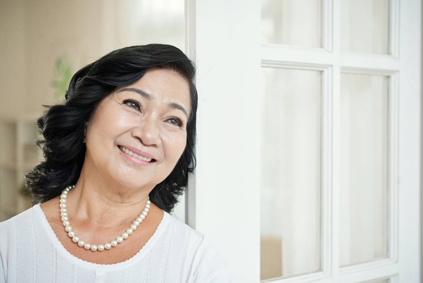 Cintura-up retrato de mulher asiática idosa atraente em colar de pérolas inclinado nas portas, olhando para o lado sonhador e sorrindo alegremente
 - Foto, Imagem