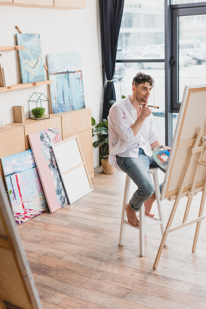 selective focus of smiling artist sitting on high chair at easel with canvas - Photo, Image