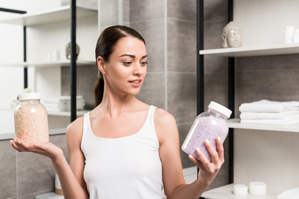 attractive brunette woman holding glass jars with sea salt in bathroom  - Zdjęcie, obraz
