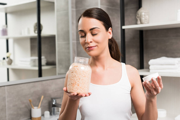 attractive brunette woman smelling sea salt while holding glass jar in bathroom  - Zdjęcie, obraz