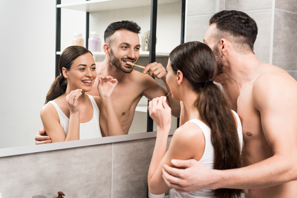 bearded boyfriend brushing teeth near girlfriend using dental floss while looking at mirror in bathroom  - Fotó, kép