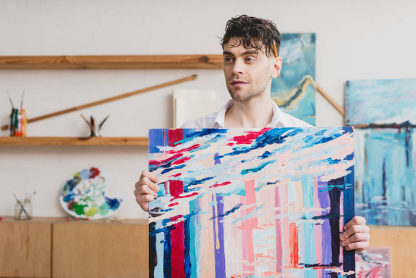 gutaussehender Künstler hält bunte abstrakte Malerei in der Hand und schaut weg - Foto, Bild