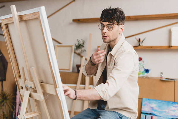 pohledný umělec v brýle sedí poblíž stojan s plátnem a při pohledu na fotoaparát - Fotografie, Obrázek
