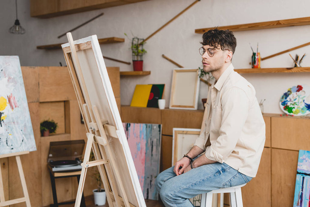 pohledný umělec v brýle sedí poblíž stojan s plátnem v malířském ateliéru - Fotografie, Obrázek