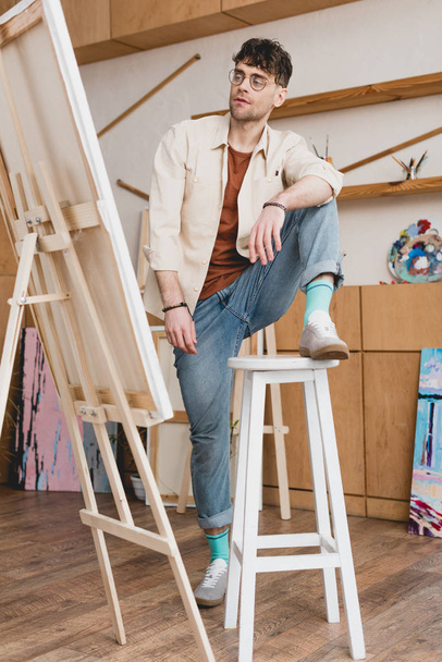 handsome artist standing with foot on chair and looking at easel with canvas - Фото, изображение