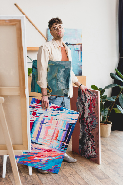 serious artist in eyeglasses holding colorful paintings and looking at camera - Photo, Image
