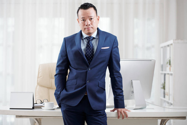 Portrait of confident Vietnamese businessman in suit leaning on his table and looking at camera  - Photo, Image