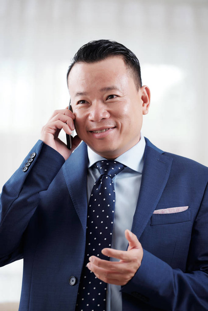 Uśmiechający się wietnamski przedsiębiorca, tłumacząc coś do partnera handlowego na telefon - Zdjęcie, obraz