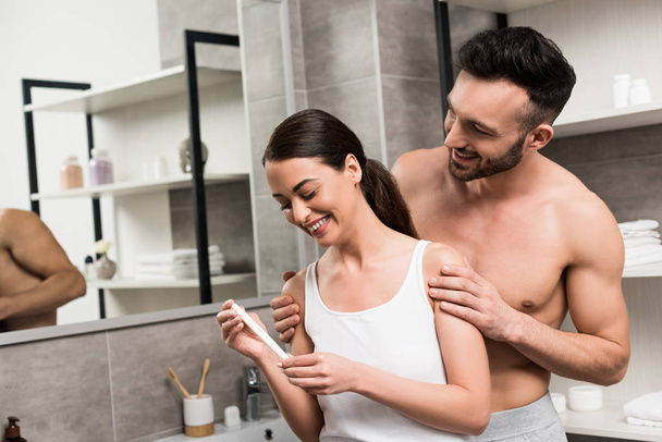 Gelukkig shirtless man knuffelen vrolijke vriendin bedrijf zwangerschapstest in badkamer  - Foto, afbeelding