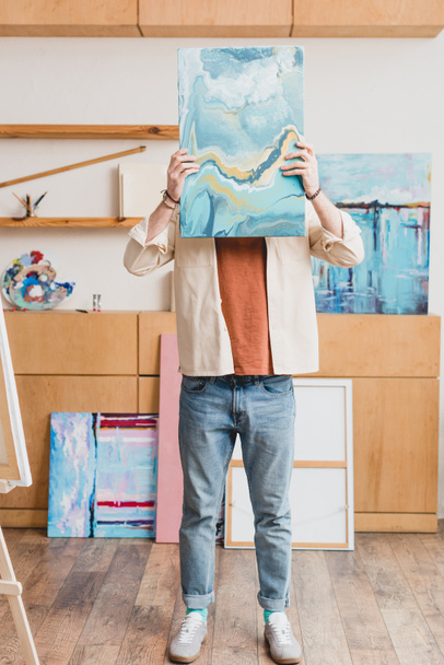 artist standing in painting studio and hiding face behind colorful abstract painting  - Foto, Bild