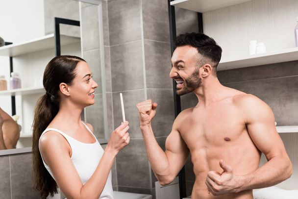 バスルームでの妊娠検査を保持している陽気なガール フレンドを見ながらジェスチャー興奮している上半身裸の男  - 写真・画像
