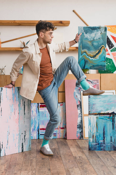 artista atractivo sosteniendo pinturas abstractas multicolores en el estudio de pintura
 - Foto, Imagen