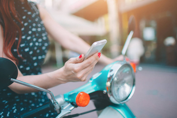 Beautiful young woman dressed casual sitting on scooter and Holdind phone. Girl sitting on the bike and using Cellular smartphone - Фото, изображение