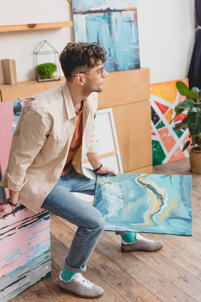 handsome artist in pink shirt and blue jeans holding colorful abstract painting and looking away - Photo, Image
