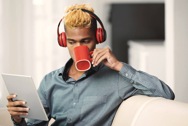 Красивий молодий чоловік в навушниках п'є каву і читає дані на планшетному комп'ютері
 - Фото, зображення