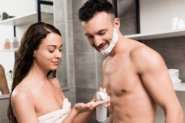 handsome bearded man with shaving foam on face looking at hand of attractive girlfriend in bathroom  - Foto, imagen
