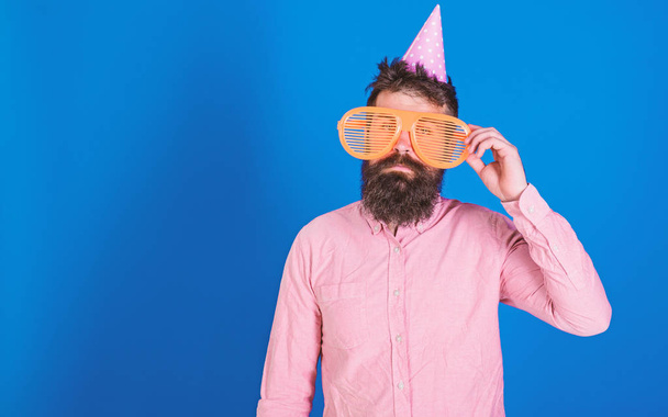 Man with beard and mustache on calm face lonely on his birthday, blue background. Guy in party hat celebrates alone. Birthday concept. Hipster in giant sunglasses celebrating birthday, copy space - 写真・画像