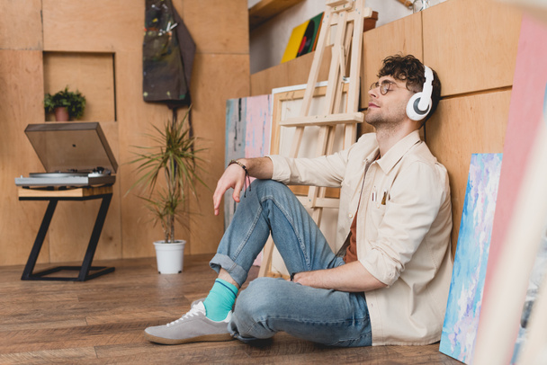 красивый художник в наушниках сидит на полу и слушает музыку с закрытыми глазами
 - Фото, изображение
