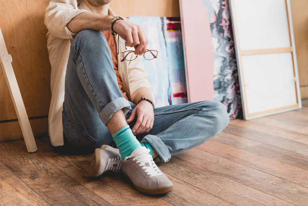 Ausgeschnittene Ansicht eines Künstlers, der auf Holzboden sitzt und eine Brille hält - Foto, Bild