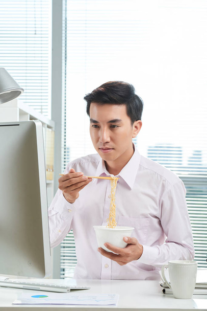 Young Vietnamese businessman reading e-mails on computer screen and eating instant noodles - Photo, Image