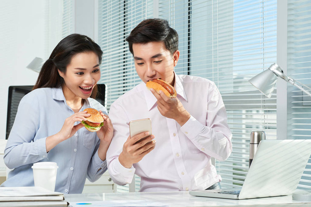 Gente de negocios comiendo comida rápida y leyendo noticias emocionantes en la pantalla del teléfono inteligente
 - Foto, Imagen