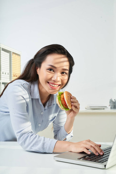 Sorridente bella donna d'affari mangiare hamburger e lavorare sul computer portatile
 - Foto, immagini